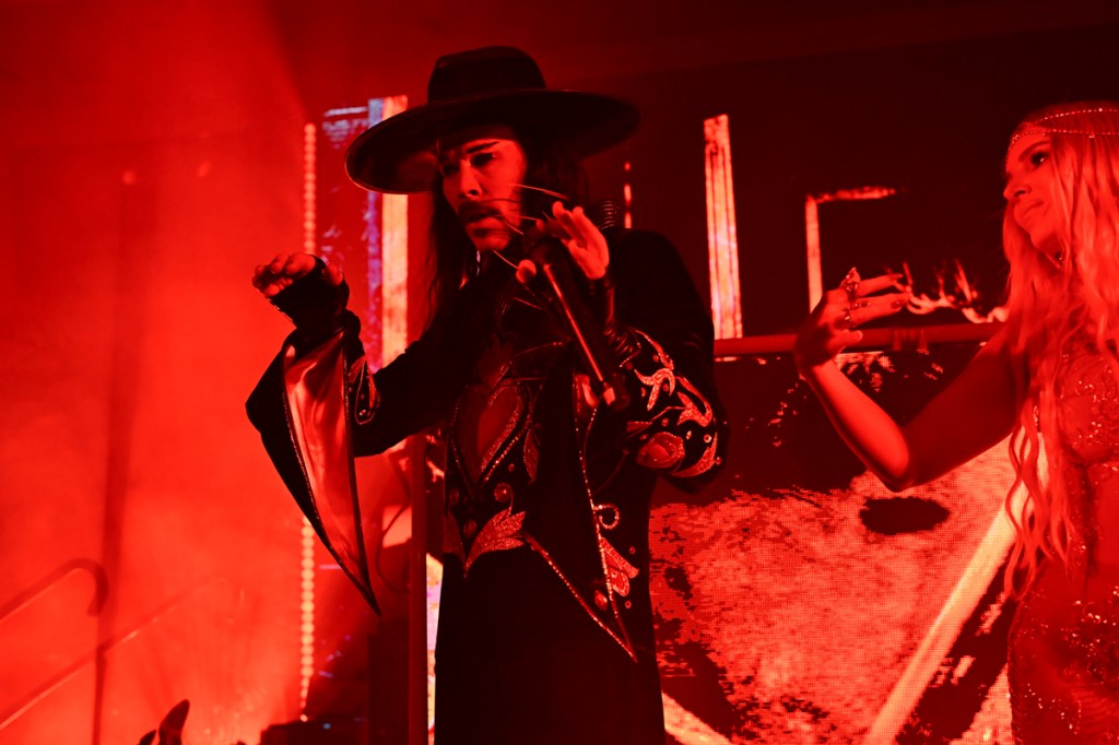 Photo of a man wearing a hat and a robe performing under red lights. 