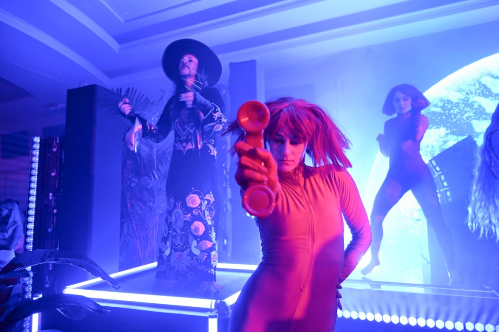 Photo of a man performing on stage and a woman wearing a wig holding a microphone. 