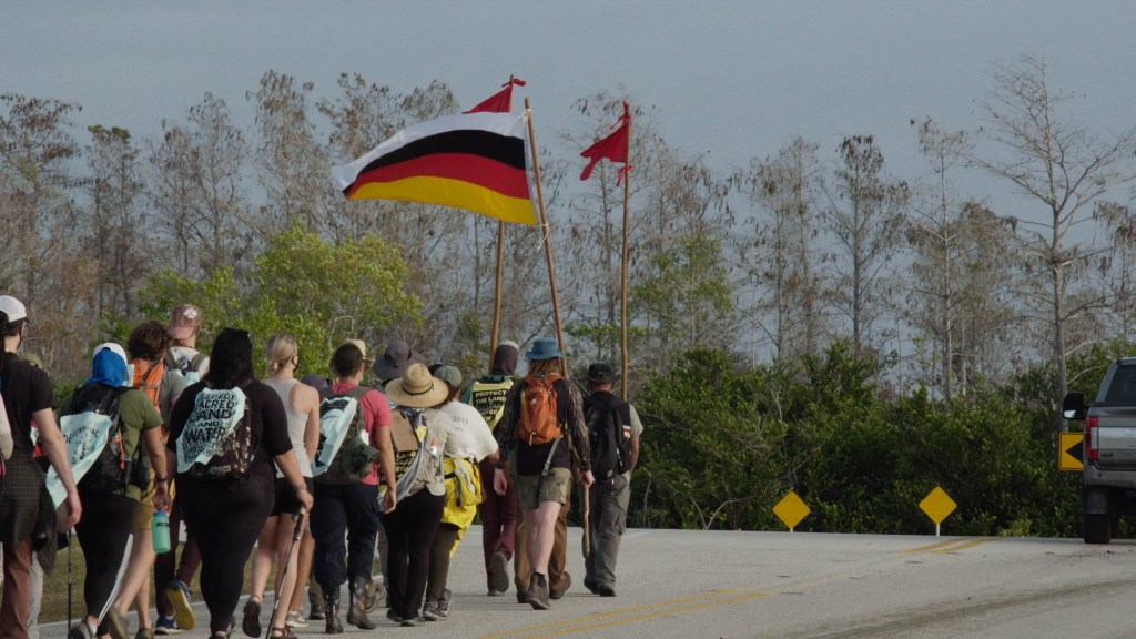 A group of people march on a dead-end street in the Everglades. One person flies the flag of the Miccosukee Tribe