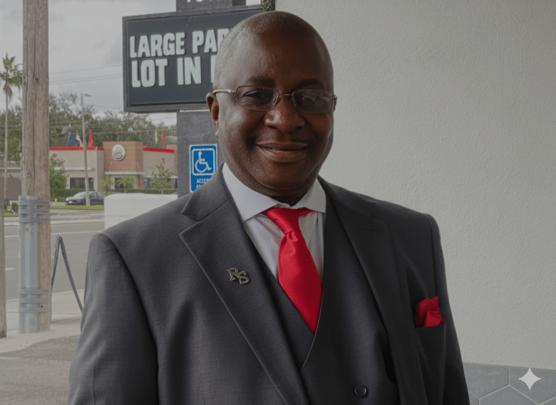 A person in a dark suit, red tie, and glasses stands outside near a wall and a sign indicating a parking lot.