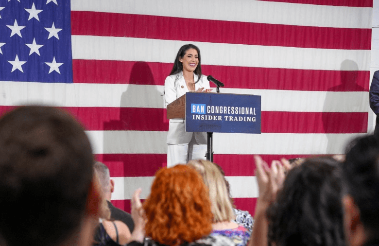 A public official in a white suit stands at a podium reading “Ban Congressional Insider Trading,” speaking to an audience in front of a large U.S. flag as people in the crowd clap in the foreground.