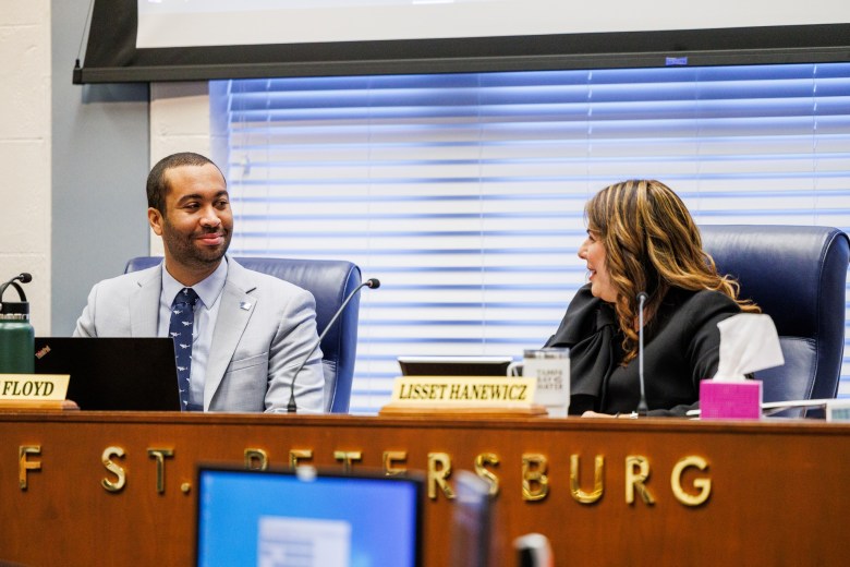 Richie Floyd and Lisset Hanewicz are pictured seated at a long wooden dais during a formal meeting of the St. Petersburg City Council. Floyd is dressed in a light grey suit with a patterned blue tie, looking toward Hanewicz.