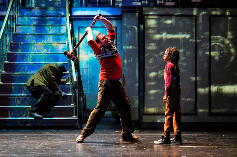 A high-tension stage scene showing a person in a red-and-white patterned sweater and brown corduroy trousers raising a large mallet high above their head, prepared to strike. They are looking down at a young child in orange overalls and a red plaid shirt who stands still and looks back at them. In the background, another figure is crouched on a blue-carpeted staircase under dim green and yellow lighting.