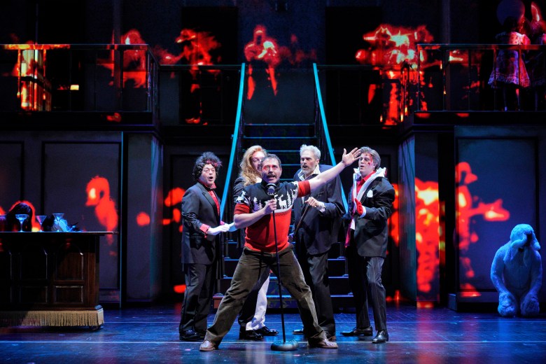 A dramatic, wide-angle stage production shot of several performers. In the center, a person in a red, black, and white holiday sweater with reindeer patterns stands behind a vintage microphone with their arm raised high. They are surrounded by four other performers in dark formal wear and theatrical makeup, one of whom has blood on their face, set against a dark stage with ghostly red and yellow projections.