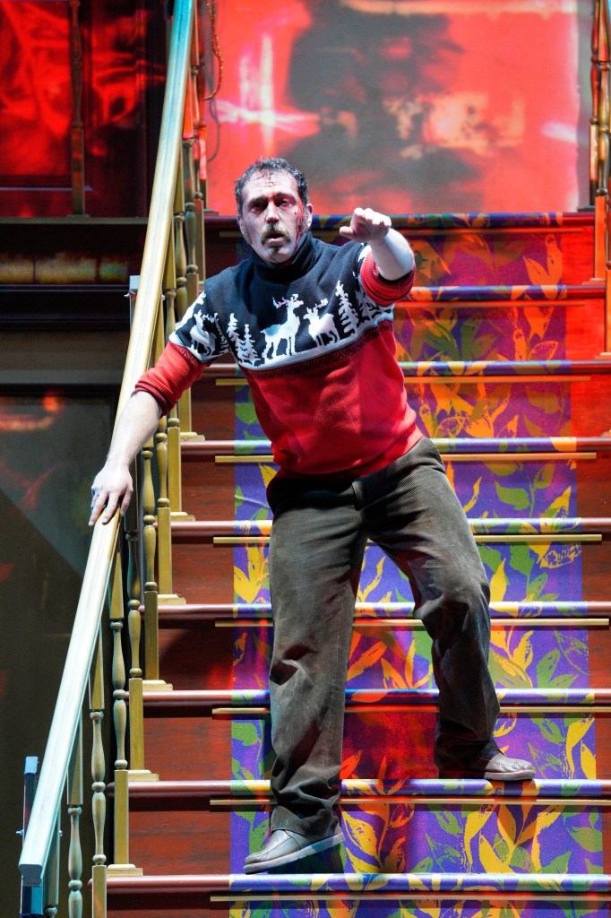 A dynamic stage shot of a person with a bloodied face descending a wooden staircase. They are wearing a red-and-white holiday-themed sweater and brown trousers, holding onto the banister with one hand while reaching out toward the viewer with the other. The staircase is covered in a vibrant purple-and-yellow floral runner, and the background features a blurry red-and-yellow projection.