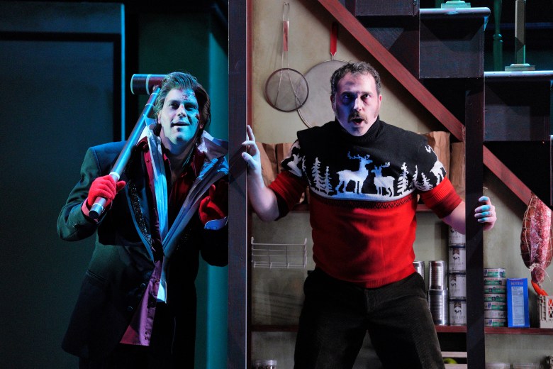 A close-up stage shot of two performers appearing from behind a wooden structure. On the right, a person in a red, black, and white reindeer-patterned sweater looks forward with a shocked or wide-eyed expression. To their left, another person in a dark suit with a bloodied face and red gloves holds a large mallet over their shoulder and looks on with a sinister smile.