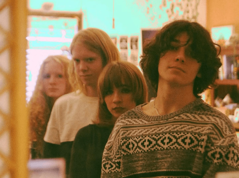 A warm-toned, slightly grainy group portrait of four people standing in a line, receding into the background. The individual in the foreground has wavy dark hair and is wearing a grey patterned knit sweater. The people behind them have varying hair lengths and colors, all looking toward the camera with neutral expressions in a dimly lit indoor setting with a soft green glow in the background.