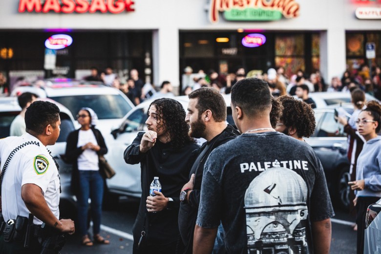 n a crowded parking lot at night, an individual with long curly hair and a black shirt speaks with a law enforcement officer wearing a white uniform and a "Hillsborough County Sheriff" patch. In the foreground, another person stands with their back to the camera wearing a grey t-shirt featuring a graphic of the Dome of the Rock and the word "PALESTINE."