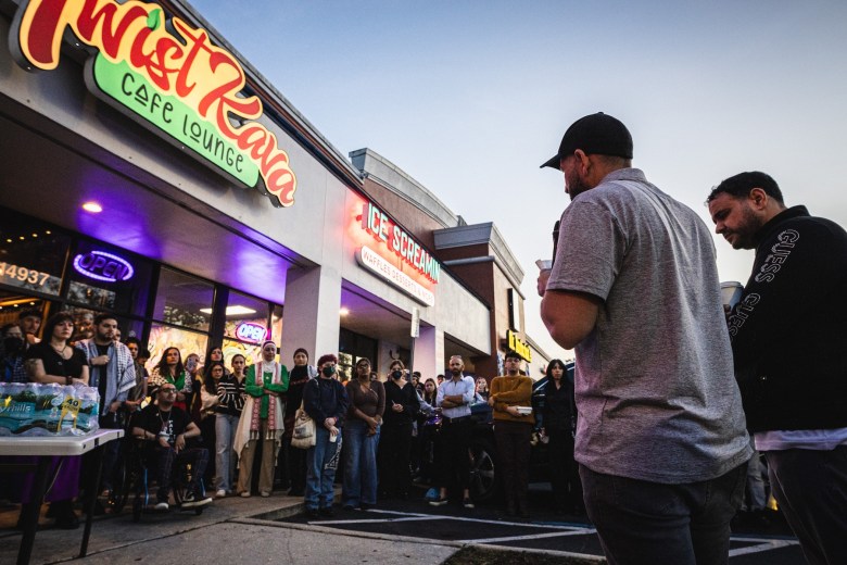 A person in a grey shirt and black cap stands with their back to the camera, addressing a diverse crowd gathered in front of businesses labeled "Twist Kava Cafe Lounge" and "Ice Screamin" at night. Several individuals in the crowd wear traditional scarves, and a table with cases of bottled water is visible in the foreground.