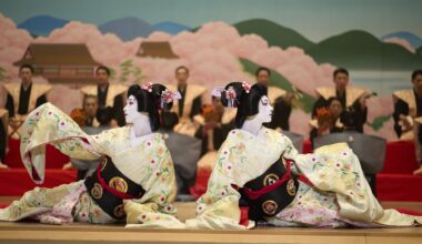 film still showing two geishas on a stage in front of men in uniforms