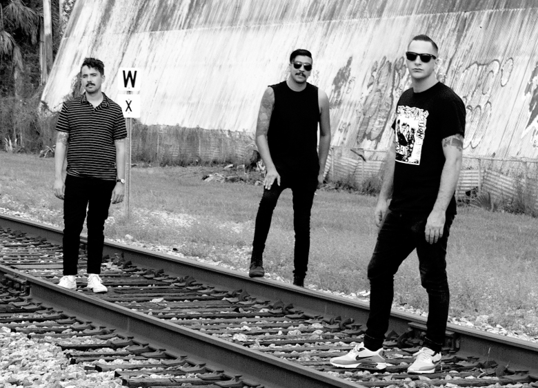 A black-and-white group portrait of three people standing on and near a set of railroad tracks. The individual on the left is wearing a striped polo shirt and dark trousers; the person in the center is wearing a sleeveless black top and sunglasses; and the person on the right is wearing a graphic t-shirt, sunglasses, and athletic sneakers. In the background, a large industrial building with graffiti and a "W X" railroad sign are visible.