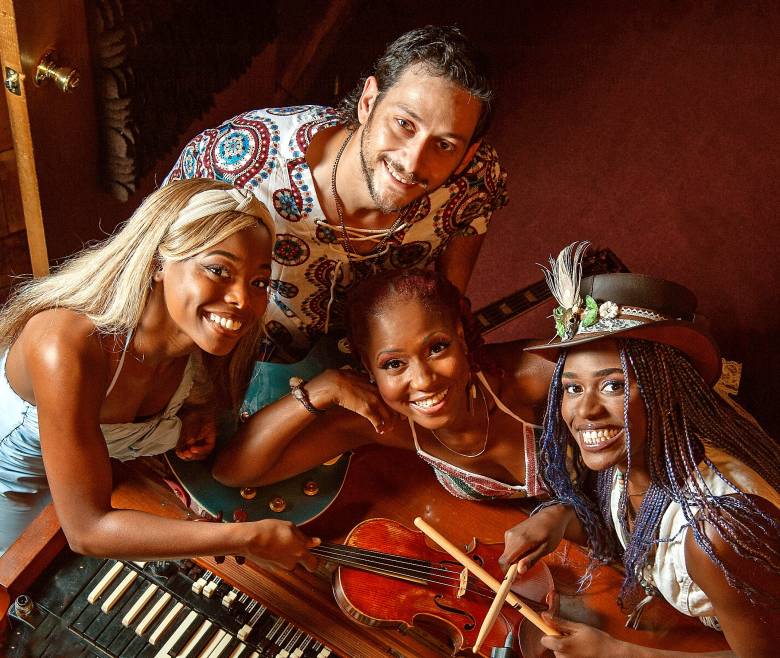 Four members of a musical group smile together for a high-angle photo. They are gathered around an organ and a violin; one person wears a patterned shirt, another has blonde hair with a headband, another has reddish hair, and the fourth wears a brown hat with a feather and has blue-braided hair.