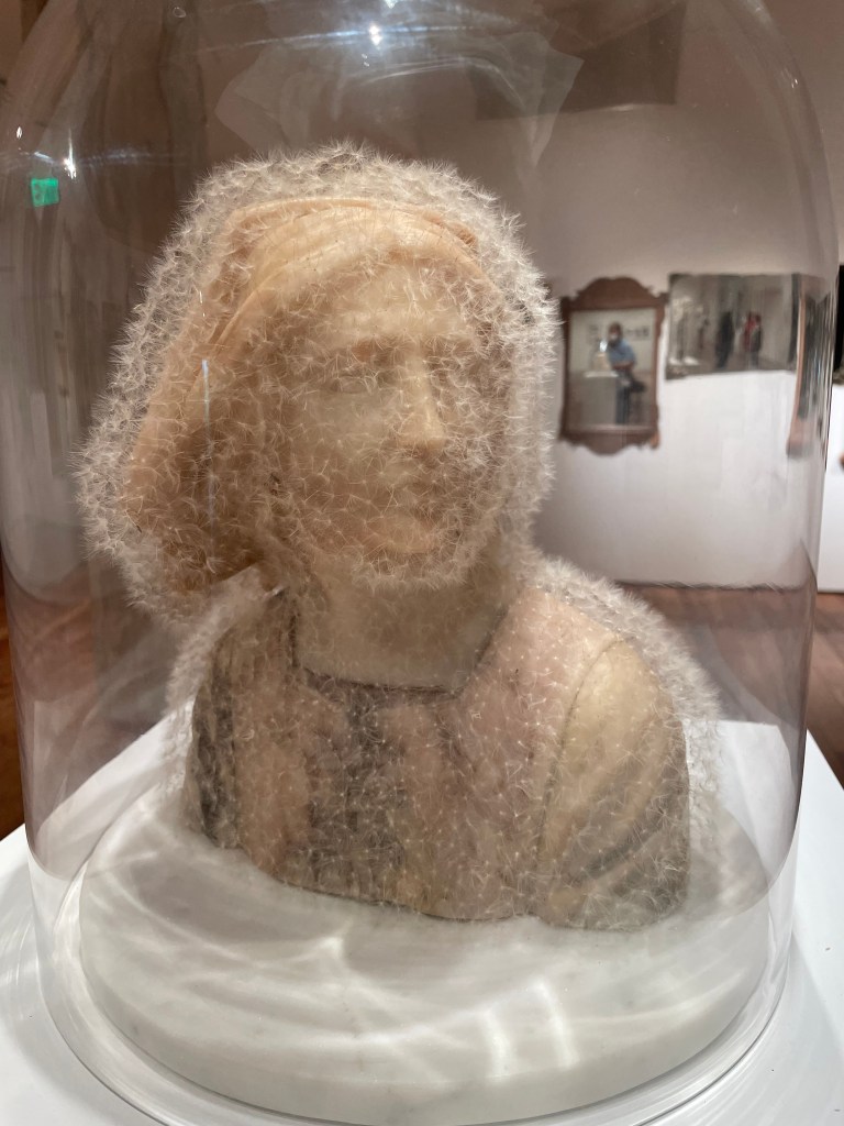 A sculpture of a person's bust, titled "Stone Joan of Arc," displayed inside a large clear glass cloche. The light-colored stone figure is entirely covered in delicate, white dandelion seeds, creating a soft, fuzzy texture over the facial features and shoulders. The piece is set on a white circular base within an art gallery setting.