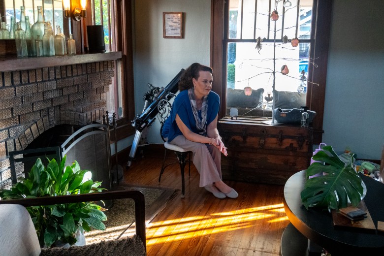 A woman sitting contemplatively on a chair in a sunlit living room with vintage decor.
