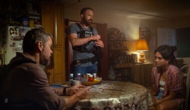 film still of two people at a dining room table speaking to a police officer