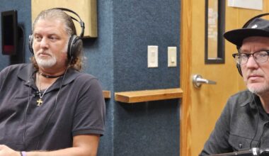 A candid shot inside a broadcast booth with sound-dampening blue and grey wall panels. Professional microphones are positioned in front of the two subjects, who are focused on a discussion off-camera.