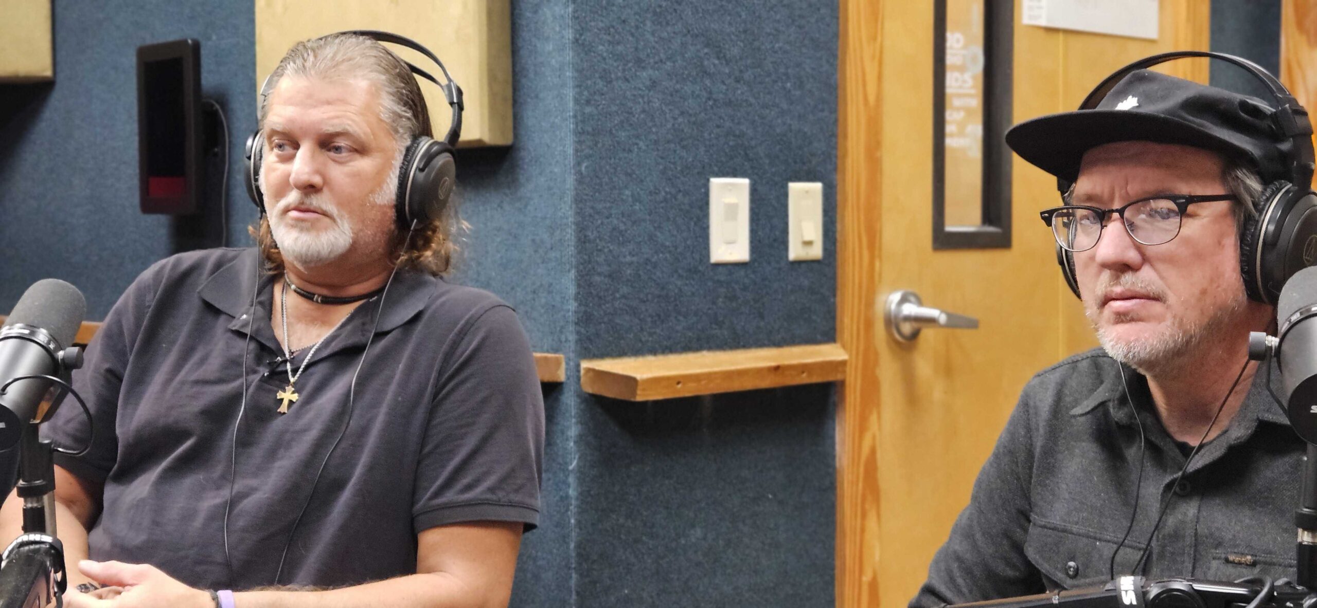 A candid shot inside a broadcast booth with sound-dampening blue and grey wall panels. Professional microphones are positioned in front of the two subjects, who are focused on a discussion off-camera.