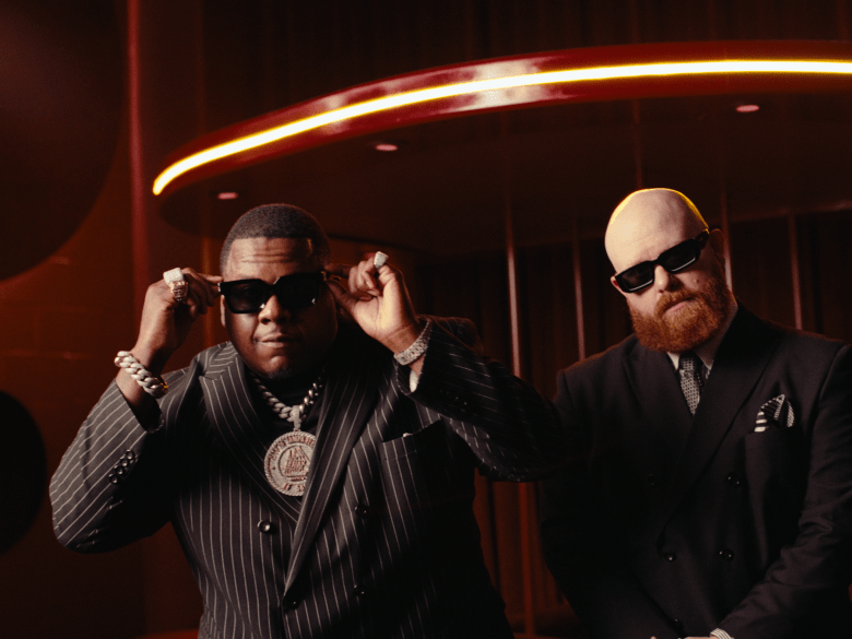 A stylish portrait of two men in dark suits and sunglasses. The man on the left wears a pinstriped suit and heavy diamond jewelry, while the man on the right has a red beard and wears a double-breasted jacket.