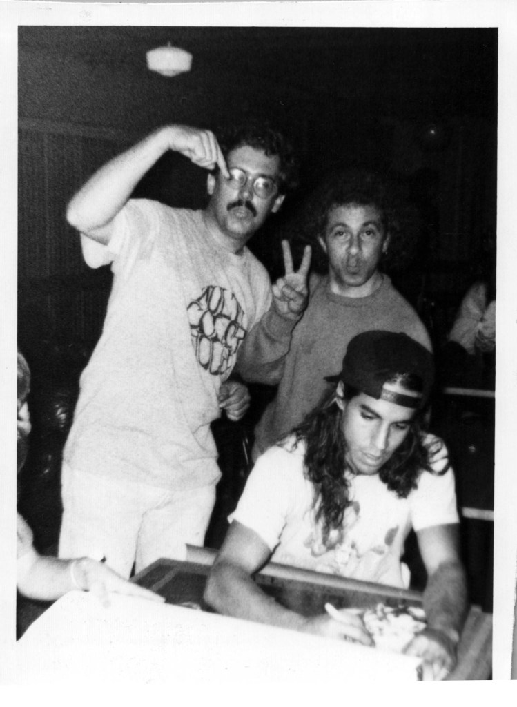 A black-and-white candid photograph from the mid-1980s featuring No Clubs Presents co-founders Tony Rifugiato (standing, top right, making a peace sign) and Dave Hundley (standing, top left, pointing to his eye) behind Anthony Kiedis of the Red Hot Chili Peppers. Kiedis is seated in the foreground, wearing a backwards baseball cap and an "Out in L.A." t-shirt, focused on signing a poster or document.