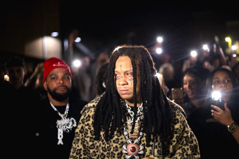 A performer with long dreadlocks and face tattoos walks through a crowd at night, wearing a leopard-print coat and layered necklaces, while people around them hold up phones with camera lights on.