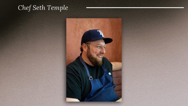 Chef Seth Temple smiles while seated inside ASH on Water Street, wearing a blue apron and Tampa Bay Rays cap.