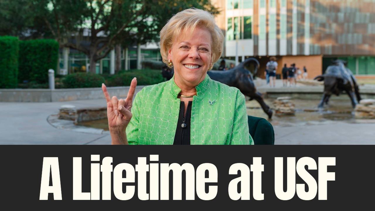 Dr. Rhea Law sits on the University of South Florida campus, smiling and flashing the Bull Horns hand sign near the USF Bull statues.