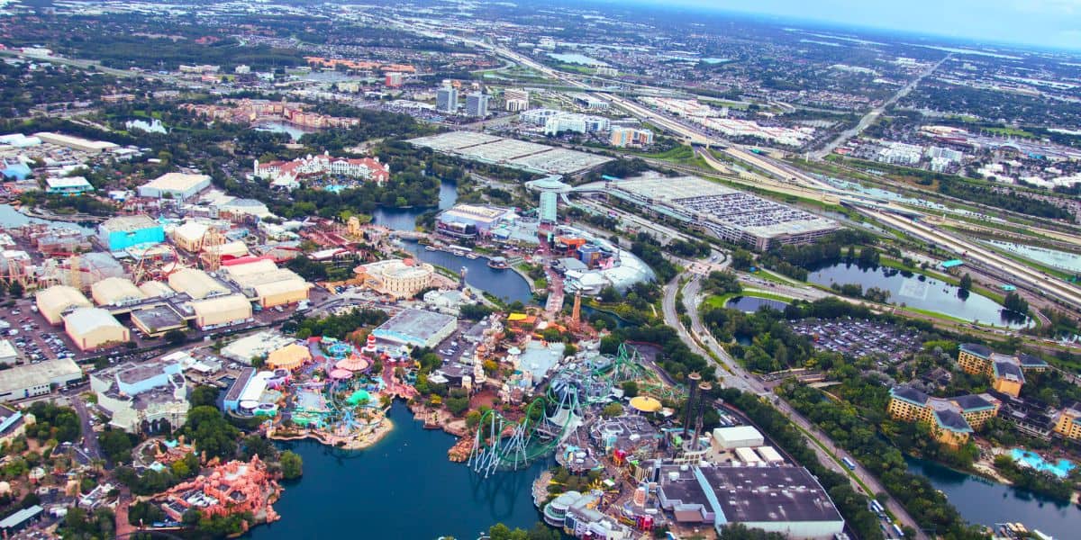 Aerial view of Universal Orlando, a large theme park featuring various attractions, rides, and roller coasters, surrounded by bodies of water. The park is adjacent to a sprawling urban area with numerous buildings, roads, and parking lots. The sky is overcast inside of Universal Studios Florida.