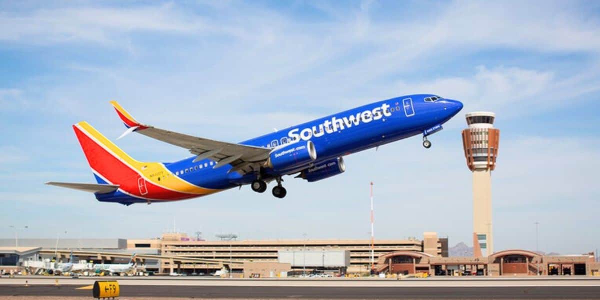 A Southwest Airlines plane with a blue fuselage and red tail is taking off from an airport runway. The airport control tower and terminal buildings are visible in the background under a clear sky, marking a history-making moment amid recent airline policy changes