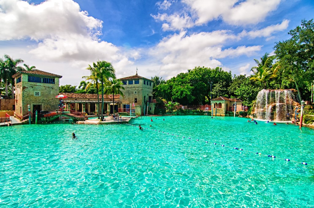 photo of a massive freshwater pool flanked by a Mediterranean Revival-style building, deck, trees, and manmade waterfall