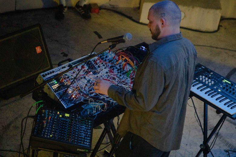 An overhead, side-profile view of a person with a shaved head and beard performing electronic music. They are standing over a complex modular synthesizer filled with a dense web of colorful patch cables, adjusting the knobs. A microphone on a stand, a small mixing board, and a secondary keyboard are also part of the setup on a concrete floor.