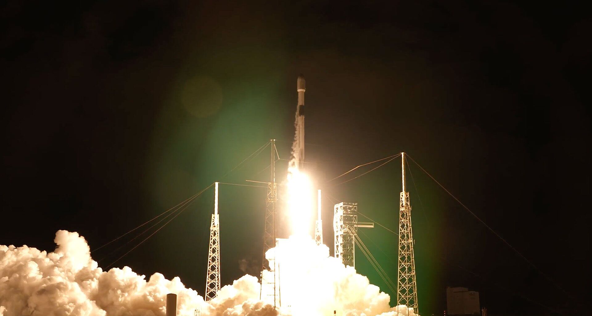 a white and black rocket lifts off into the night sky, its bright yellow-white plume lighting its launch pad below.