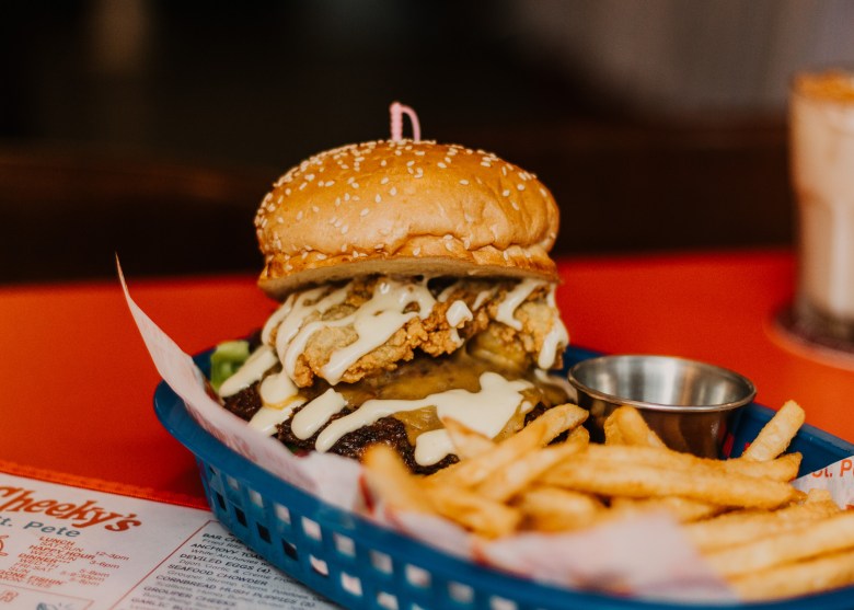 A stacked oyster burger topped with creamy sauce sits on a sesame-seed bun in a blue basket, served with fries and a small metal cup of dipping sauce on a red tabletop.