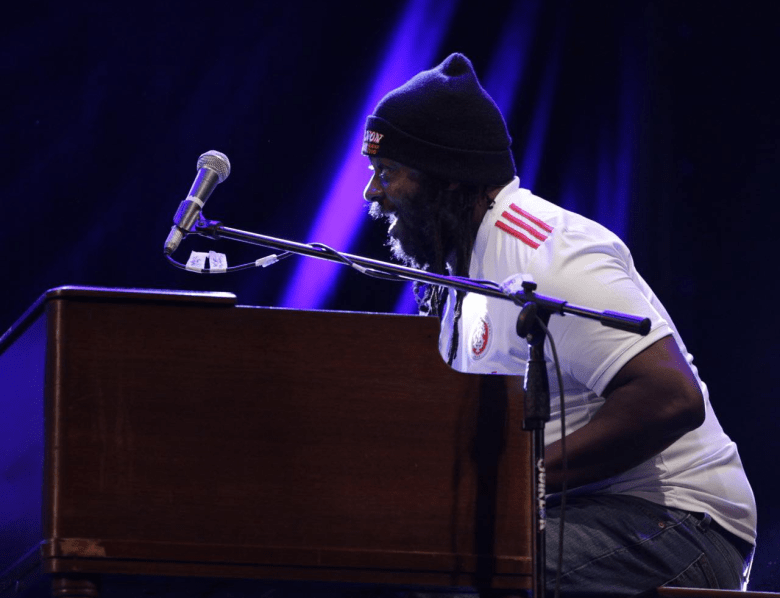 A person with a beard and long hair under a black beanie performs enthusiastically at an organ. They are wearing a white short-sleeved jersey with red stripes on the shoulders and are captured in profile, leaning toward a microphone under dramatic blue stage lighting.