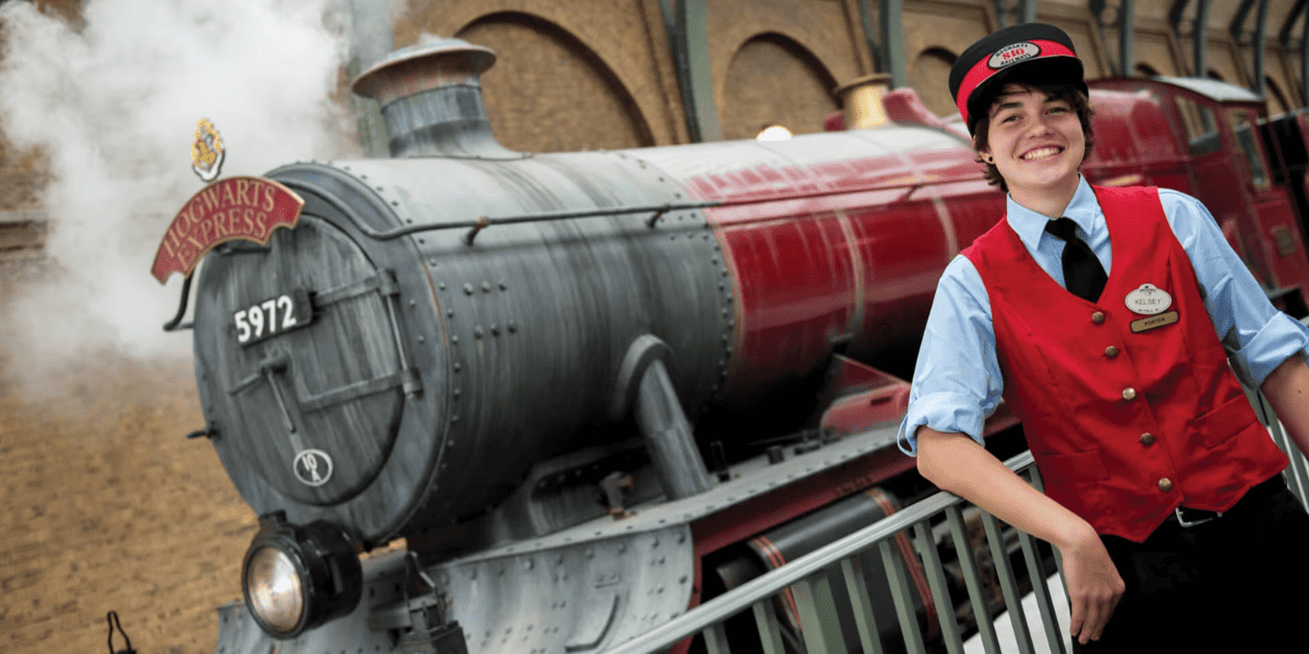 A conductor in a red vest smiles in front of the Hogwarts Express at Universal Orlando.