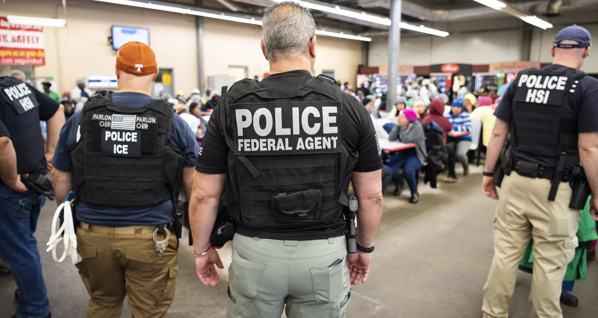 Federal agents with their back towards the camera. They wear bulletproof vests that say "Police Federal Agent" and are looking at detained migrants In the background.