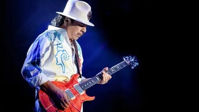 A guitarist wearing a white hat and embroidered jacket plays a red electric guitar on a dark stage illuminated by blue light.