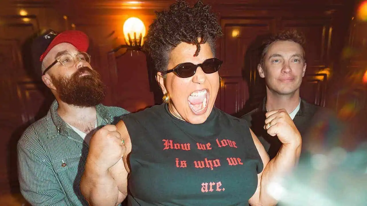 Three people pose energetically in a warmly lit room, with the person in front wearing sunglasses and a sleeveless shirt while raising clenched fists and shouting as two others stand behind them.