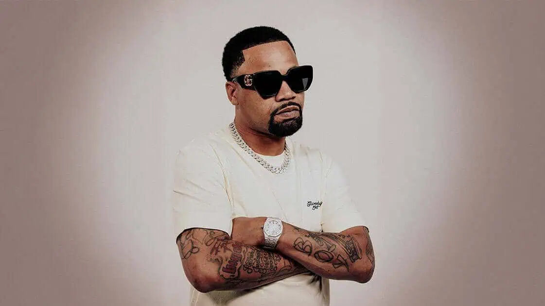 A man wearing dark sunglasses and a cream T-shirt stands with his arms crossed, showing tattooed forearms against a neutral studio background.