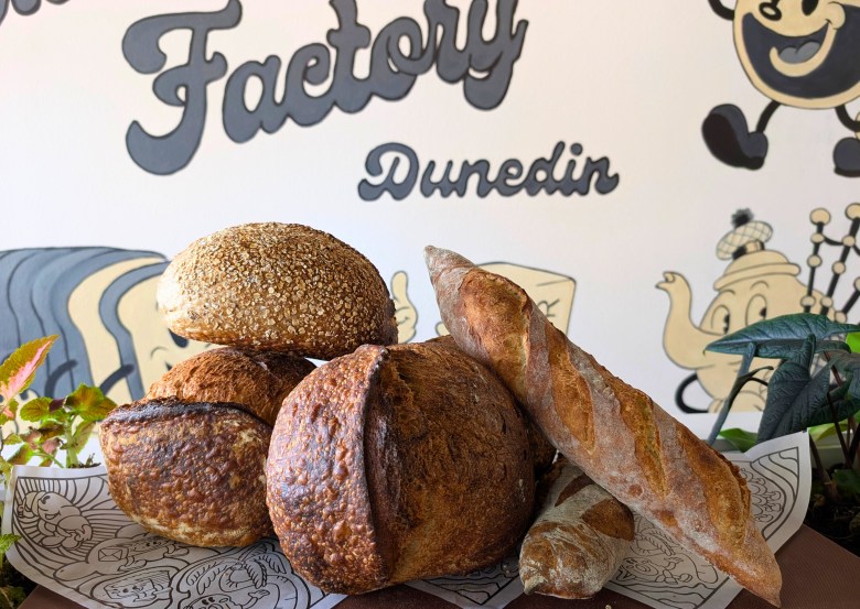 A stack of crusty artisan bread loaves and baguettes displayed in front of a mural that reads 'Factory Dunedin'