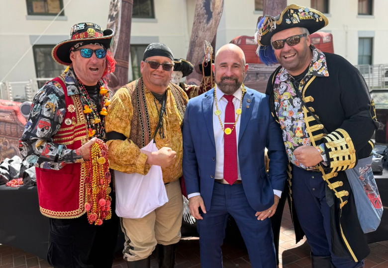 Five adults pose together outdoors at a festival or parade, smiling toward the camera. Four wear colorful pirate-style costumes with hats, beads, and decorative jackets, while one stands at center in a blue suit with a red tie. Buildings and trees are visible in the background, suggesting a lively street-festival setting.