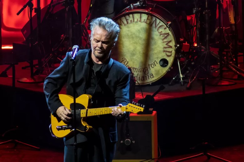 A wide shot of American singer-songwriter John Mellencamp performing live on a dimly lit stage with red backlighting. He is an older man with silver, swept-back hair, wearing a dark suit. He is playing a butterscotch blonde Fender Telecaster electric guitar and standing in front of a microphone. In the background, a large bass drum is visible, featuring his name "MELLENCAMP" and his hometown "Bloomington, Indiana" printed on the drumhead.