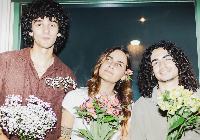 A flash-lit group portrait of three people holding bouquets of flowers. The individual on the left has voluminous curly dark hair and is wearing a brown textured button-down shirt with baby's breath tucked into the pocket. The person in the center has long wavy hair with a pink flower tucked behind their ear, looking upward. The person on the right has long curly dark hair and a wide smile, holding a bouquet of yellow and pink lilies.