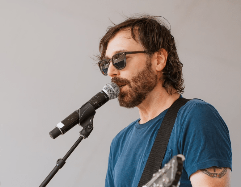 A profile shot of musician Matt Pond singing into a microphone. They have brown hair and a beard, wearing dark sunglasses and a blue t-shirt. A guitar strap is visible over their shoulder, and a small tattoo can be seen on their upper left arm against a plain light grey background.