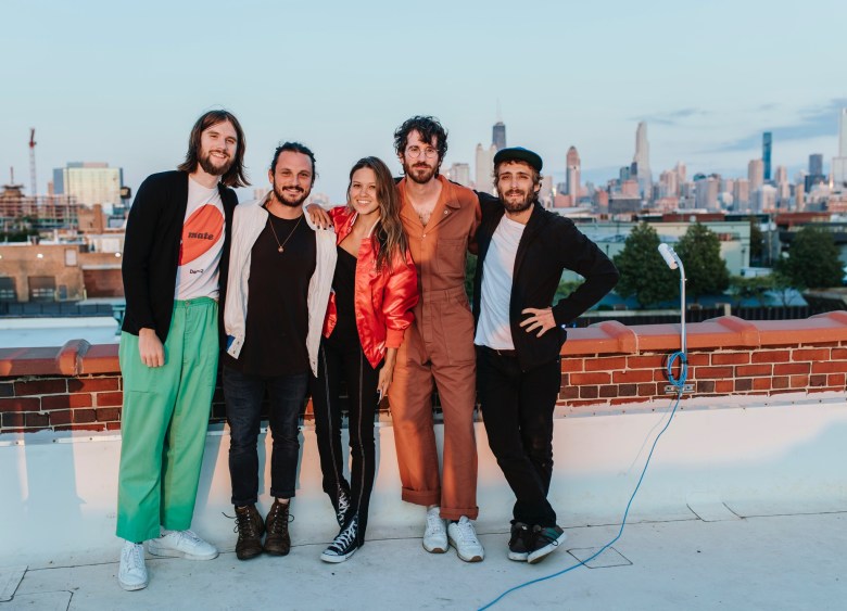 Five individuals stand together with their arms around each other on a brick-lined rooftop during sunset. The group is dressed in a mix of casual styles, including green trousers, a red satin jacket, and a copper-colored jumpsuit. In the background, a sprawling city skyline featuring several skyscrapers is visible under a clear, pale blue sky.