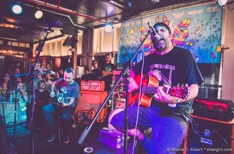A musician with dark hair and a beard sings passionately into a microphone while playing an acoustic guitar during a live indoor performance. They are wearing a black graphic t-shirt and blue shorts. In the background, another individual plays acoustic guitar while seated, and a colorful banner with cartoon illustrations hangs behind the stage area.