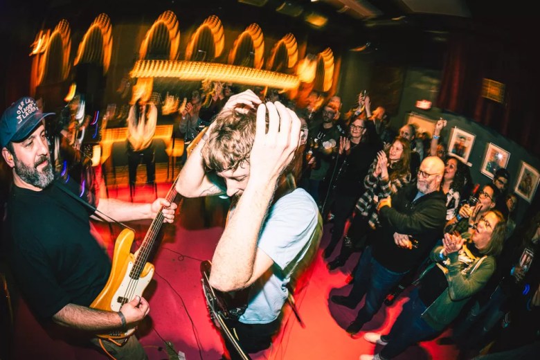 A high-energy, wide-angle live music photograph taken from a low perspective. In the foreground, a person in a black t-shirt and baseball cap plays a light-colored bass guitar while another person in a light blue shirt stands with their hands on their head. A diverse crowd in a dimly lit venue is visible in the background, clapping and cheering under warm, streaking stage lights that create a sense of motion.
