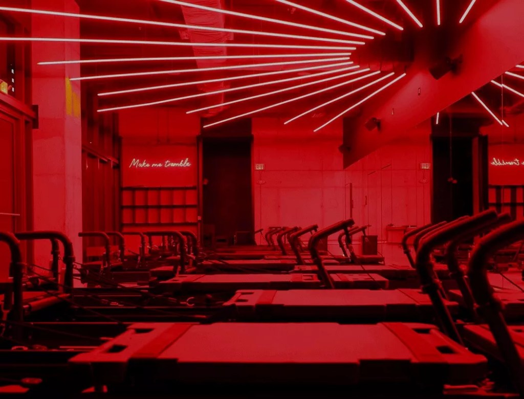 A room of Pilates reformers lit in red lighting. A neon sign on the back wall reads, "Make me tremble"