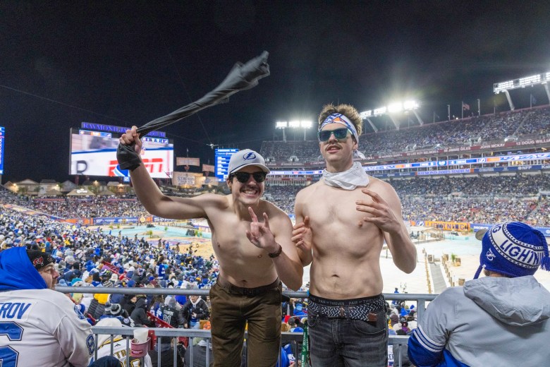 Two shirtless people celebrate in the stands of a large, crowded outdoor stadium at night. The individual on the left wears a white Tampa Bay Lightning baseball cap and sunglasses, holding up a black shirt and making a "rock on" hand gesture. The individual on the right wears a white bandana around their neck, a patterned headband, and teal-framed sunglasses. In the background, thousands of fans fill the stadium seats under bright arena lights, with a large video scoreboard visible in the distance.