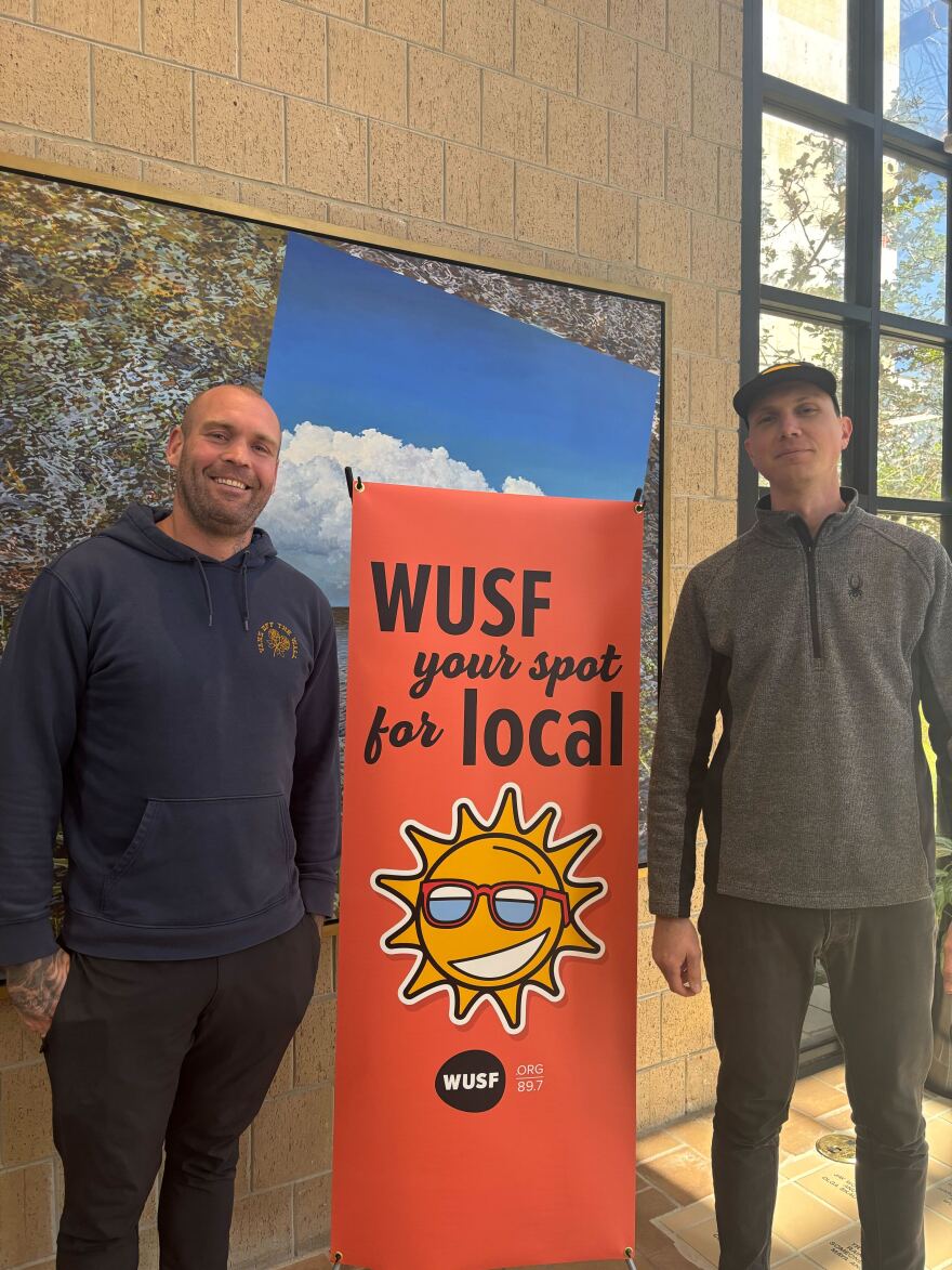 Two men stand next to a pink sign that says "WUSF your spot for local"