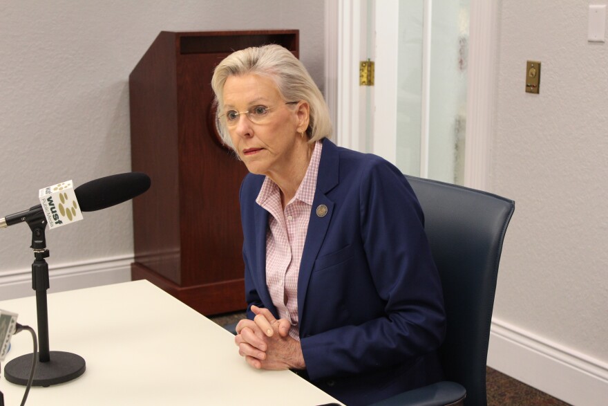 A person with blonde hair and wearing a blue blazer listens with clasped hands while seated at a desk with a microphone